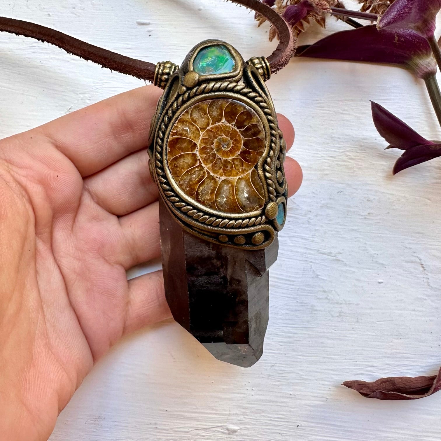 Ammonite fossil pendant on a leather cord held by a hand, with a white background.
