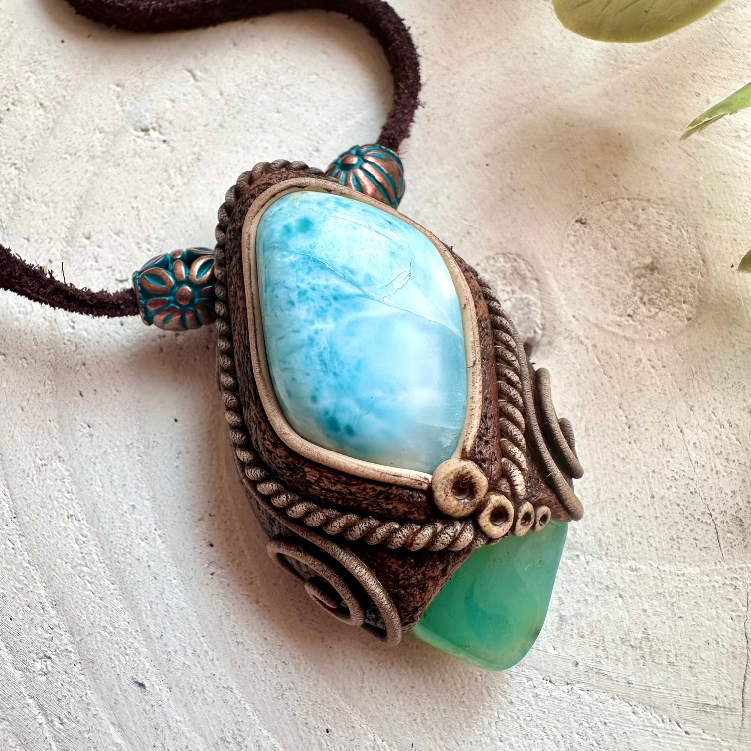 A handmade bohemian-style pendant necklace featuring a light blue marbled larimar stone, a green crysoprase stone, and clay on a dark brown cord.