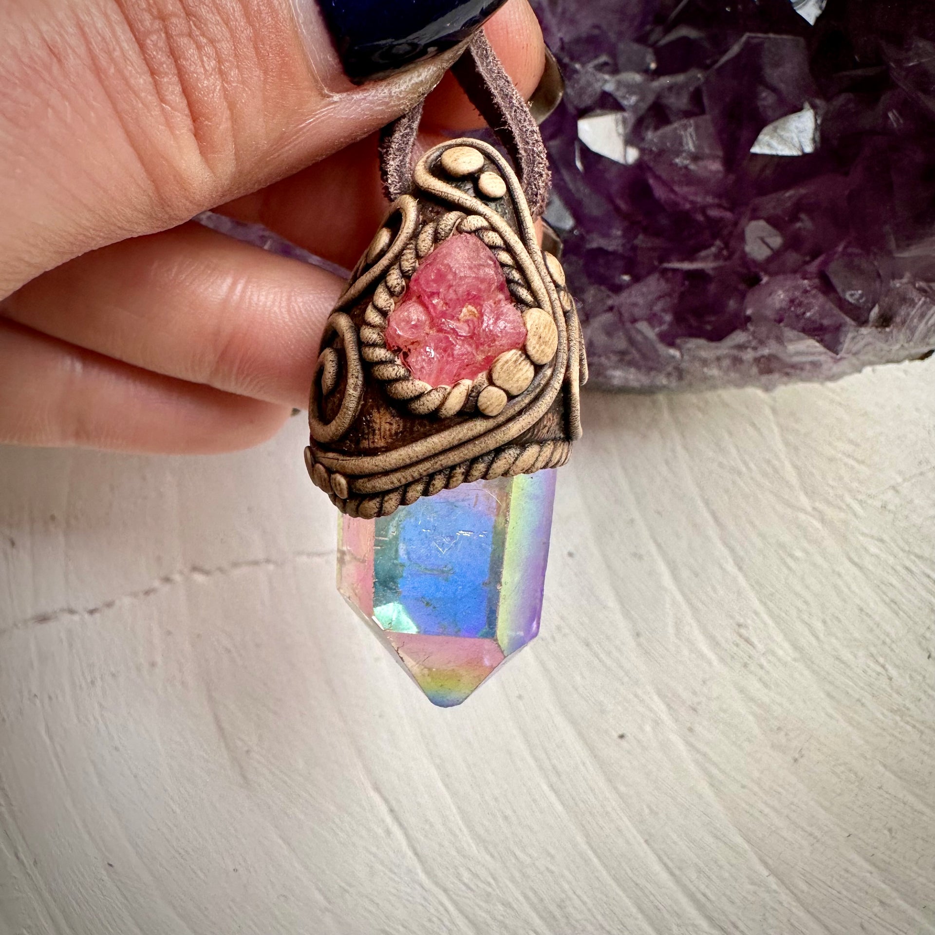 Hand holding Angel Aura Quartz and Spinel necklace with clay pendant and adjustable leather strap on a textured background.