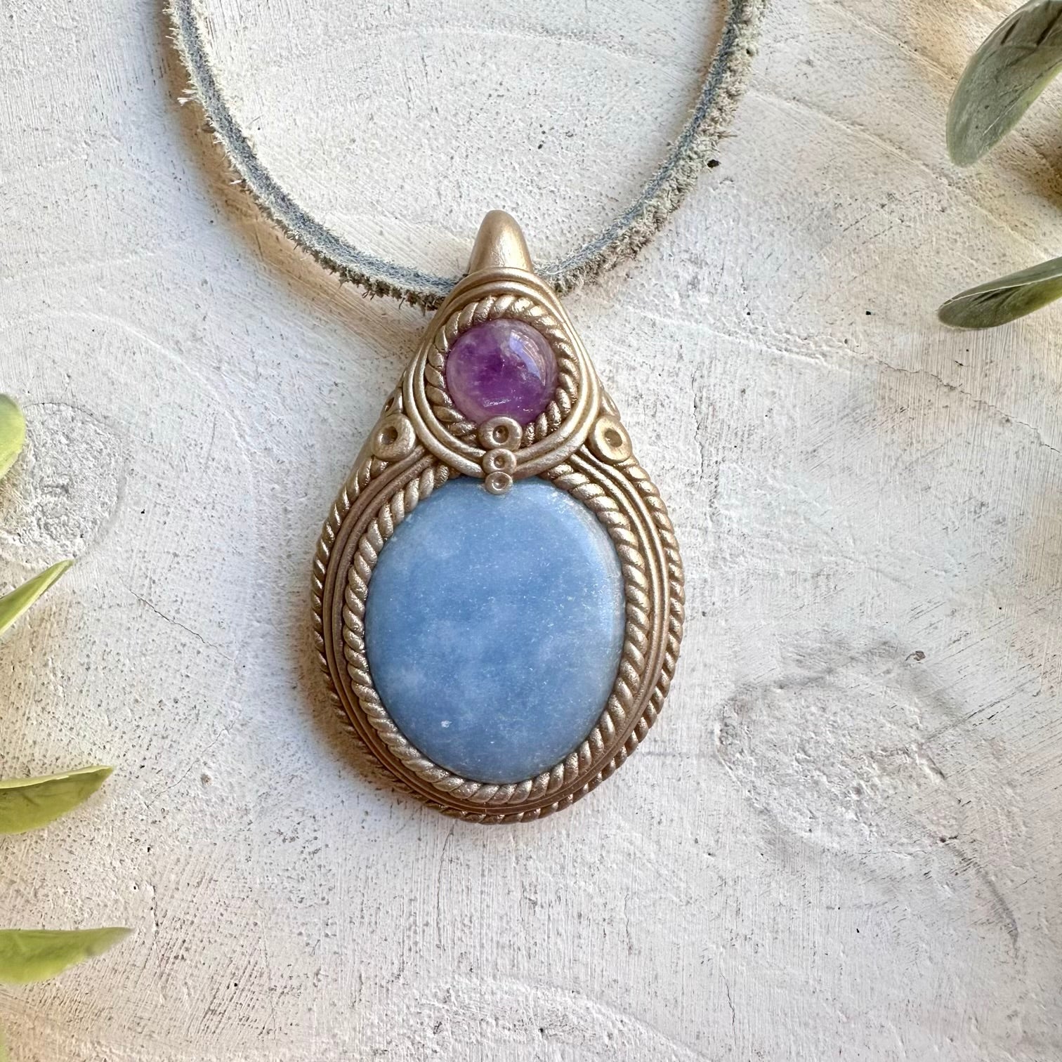 A teardrop-shaped pendant necklace featuring a large light blue stone and a smaller purple stone, set in a textured bronze-toned metal frame with a rope-like border, resting on a light textured surface with green leaves in the corners.