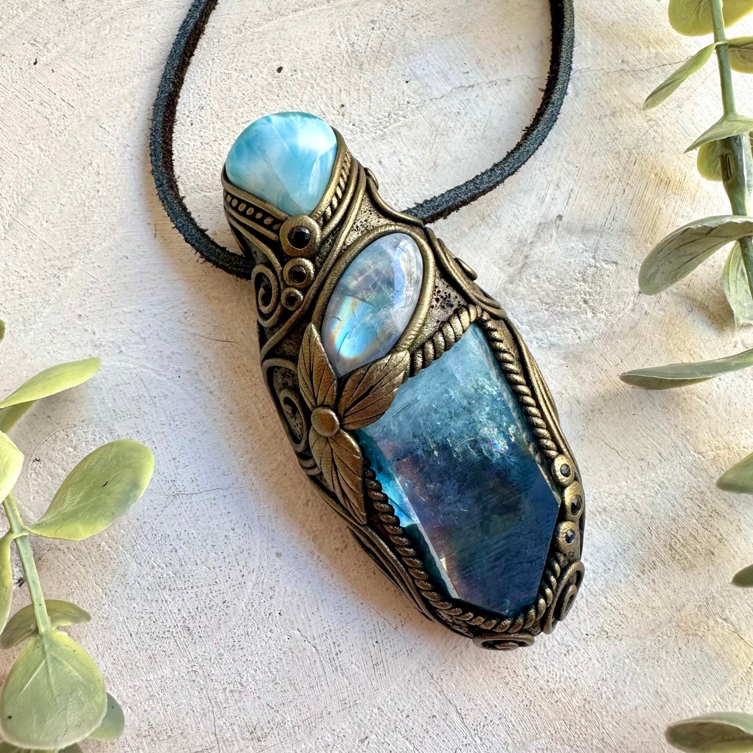 A bohemian-style pendant necklace featuring a large blue labradorite stone, a smaller iridescent blue stone, and a light blue stone, all set in an ornate bronze-toned clay frame with a black cord. The pendant is displayed on a textured beige surface with decorative green leaves in the corners.