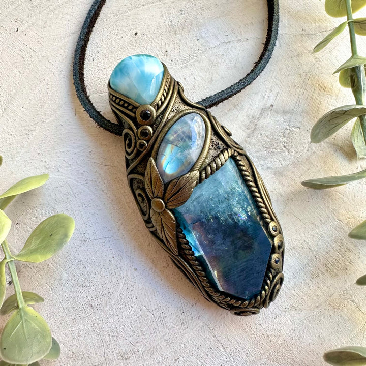 A bohemian-style pendant necklace featuring a large blue labradorite stone, a smaller iridescent blue stone, and a light blue stone, all set in an ornate bronze-toned clay frame with a black cord. The pendant is displayed on a textured beige surface with decorative green leaves in the corners.