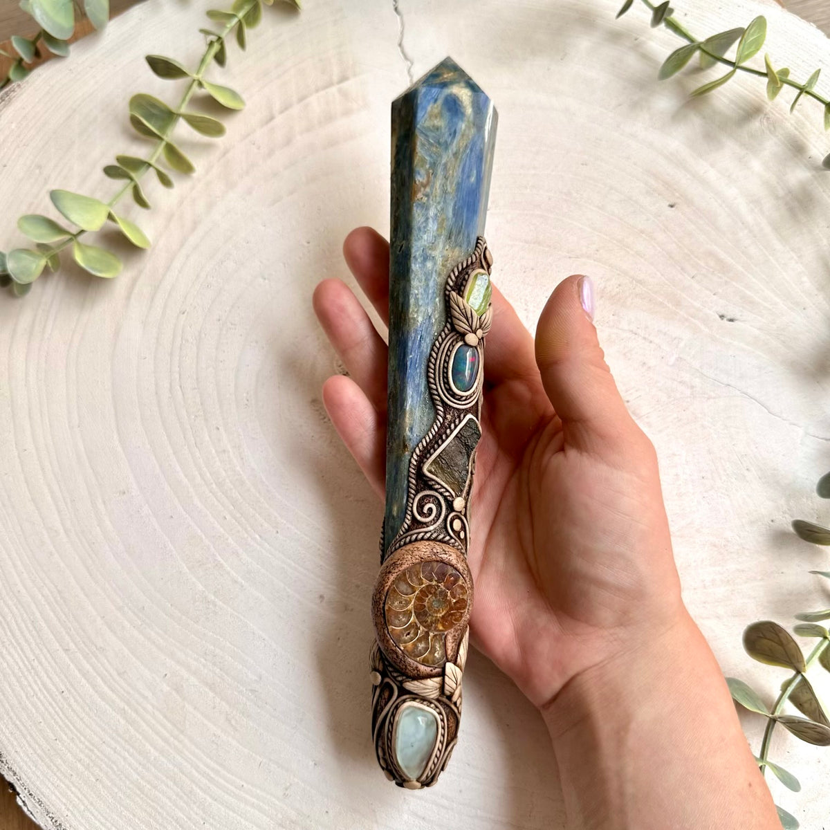 Handmade Blue Kyanite crystal wand with Ammonite, Aquamarine, Raw Moldavite, Black Ethiopian Opal and Peridot, a one of a kind spiritual crystal tool combining deep blue Kyanite with an ancient Ammonite fossil and a striking collection of natural gemstones. This unique healing crystal wand is inspired by traditional associations of clarity, communication, grounding, calmness, intuition, transformation, renewal and spiritual growth.