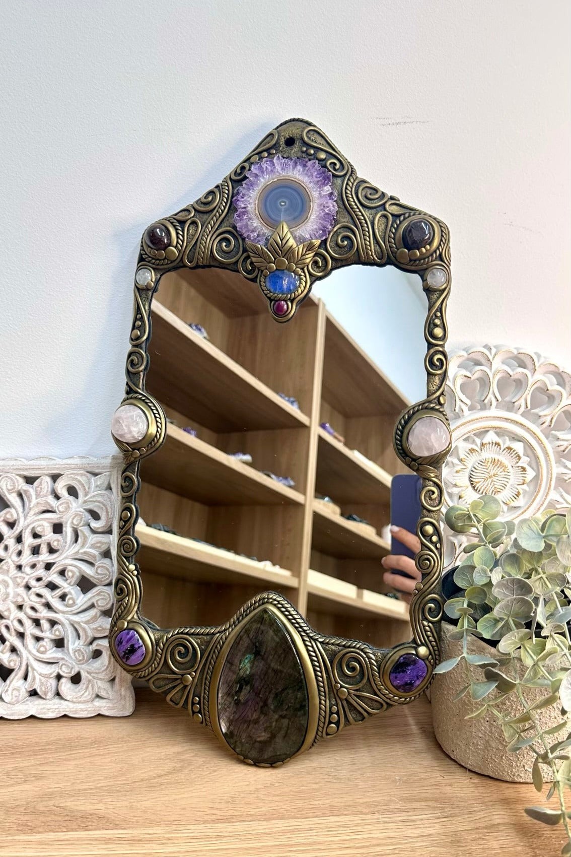 An ornate decorative wall mirror with an antique bronze frame, featuring carved scrollwork and large purple and blue gemstone accents. The mirror is shown resting on a light wood surface, reflecting a wooden shelving unit in the background, with a white carved wall panel and a potted eucalyptus plant on either side.