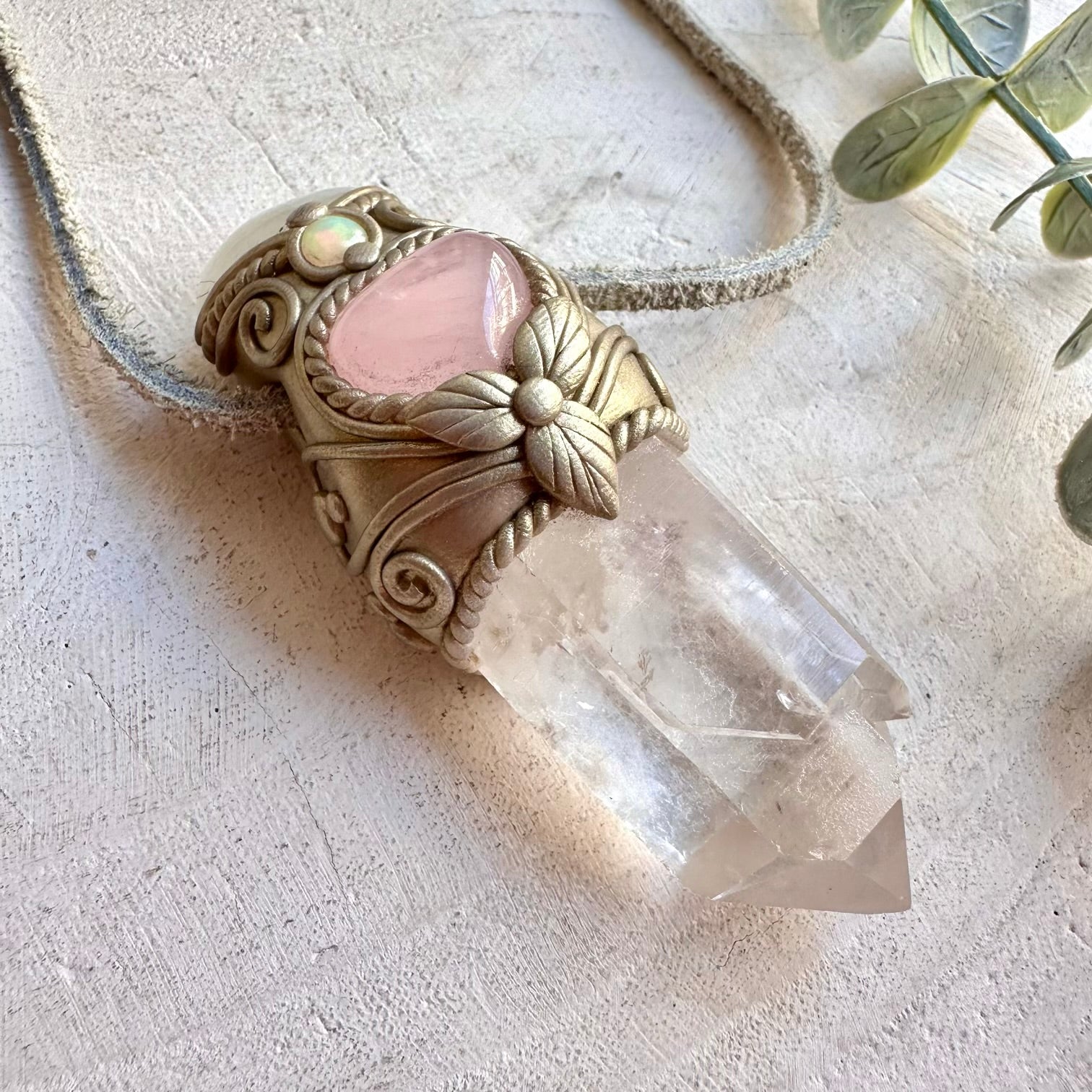 A handmade pendant necklace featuring a clear quartz crystal point set in an ornate gold-toned filigree frame with a pink heart-shaped inlay, resting on a textured beige surface with a rope cord.