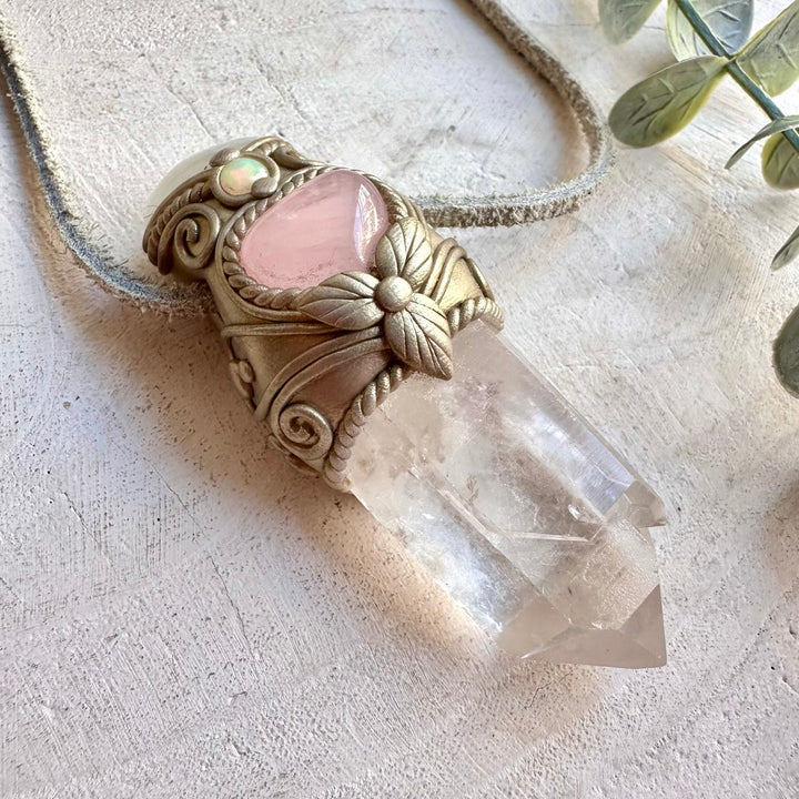 A handmade pendant necklace featuring a clear quartz crystal point set in an ornate gold-toned filigree frame with a pink heart-shaped inlay, resting on a textured beige surface with a rope cord.